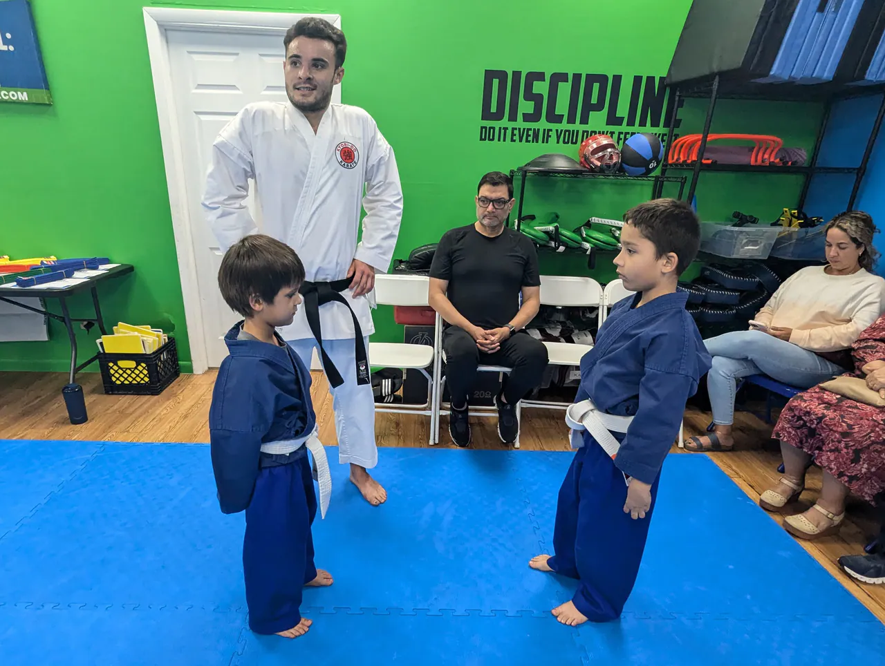 Two young beginners in blue karate uniforms facing each other while an instructor and parents watch